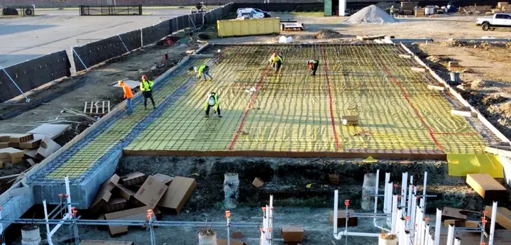 A crew in hard hats and high-visibility vests tying rebar over a new foundation, with plumbing rough-in below